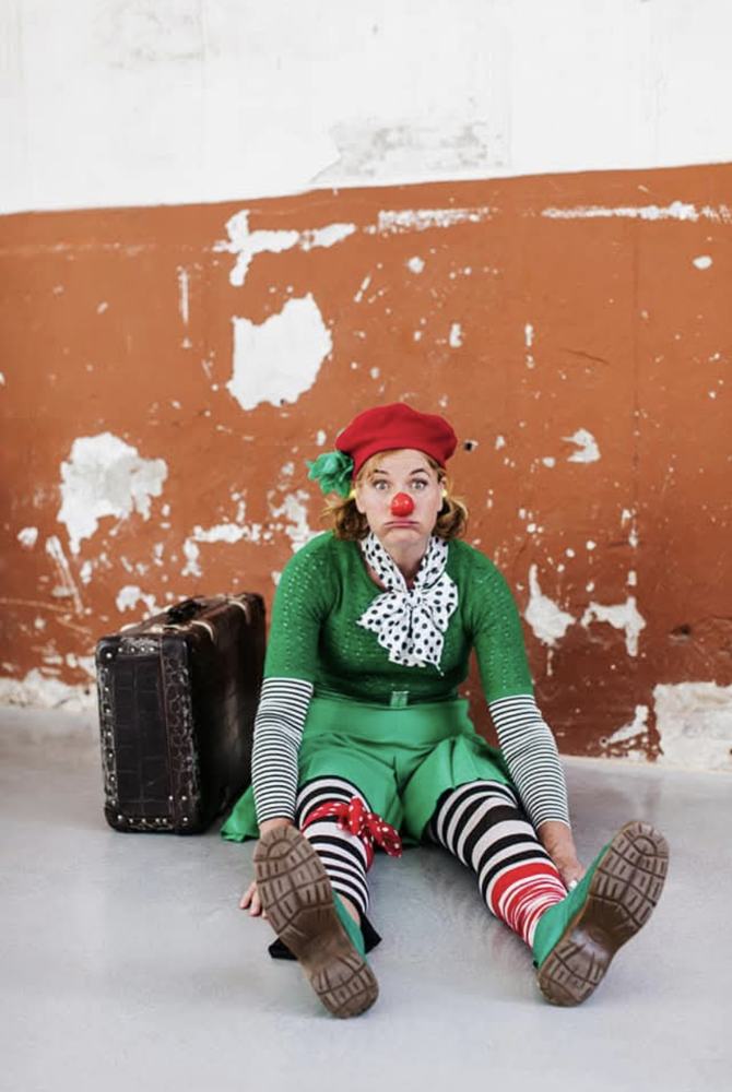 Clown Pip zit op de grond naast haar koffer, voor een oranje muur